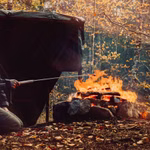 Eine Person kniet an einem Lagerfeuer im Wald und röstet Essen mit dem Stabilotherm Teleskop Grillstab. Das Feuer glüht zwischen Felsen Herbstblätter und Bäume umgeben sie und eine dunkle Plane oder ein Zelt steht in der Nähe.