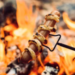 Der Stabilotherm Teleskop Grillstab für das Lagerfeuer hält einen in Teig eingewickelten Hot Dog über ein Lagerfeuer mit leuchtend orangefarbenen Flammen und Glut im Hintergrund.