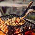 Stabilotherm Pfanne für das Lagerfeuer mit Klappgriff_31901 Eine behandschuhte Hand rührt Tortellini und Gemüse in einer Stabilotherm Pfanne für das Lagerfeuer mit Klappgriff über einer offenen Flamme auf einem Metallgrill im Freien.