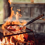 Stabilotherm Pfanne für das Lagerfeuer mit Klappgriff_31902 Die Stabilotherm Pfanne für das Lagerfeuer mit Klappgriff sitzt auf einem Grillrost über einem Holzfeuer gefüllt mit geschnittenem Gemüse. Ein Metallspatel ruht in der Pfanne die vor einem rustikalen Holzhintergrund steht.