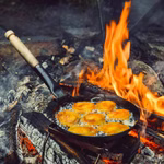 Stabilotherm Pfanne für das Lagerfeuer mit Klappgriff_31904 Die Stabilotherm Pfanne für das Lagerfeuer mit Klappgriff brät goldbraune Speisen über lodernden Flammen und brennenden Holzscheiten am offenen Feuer im Freien.