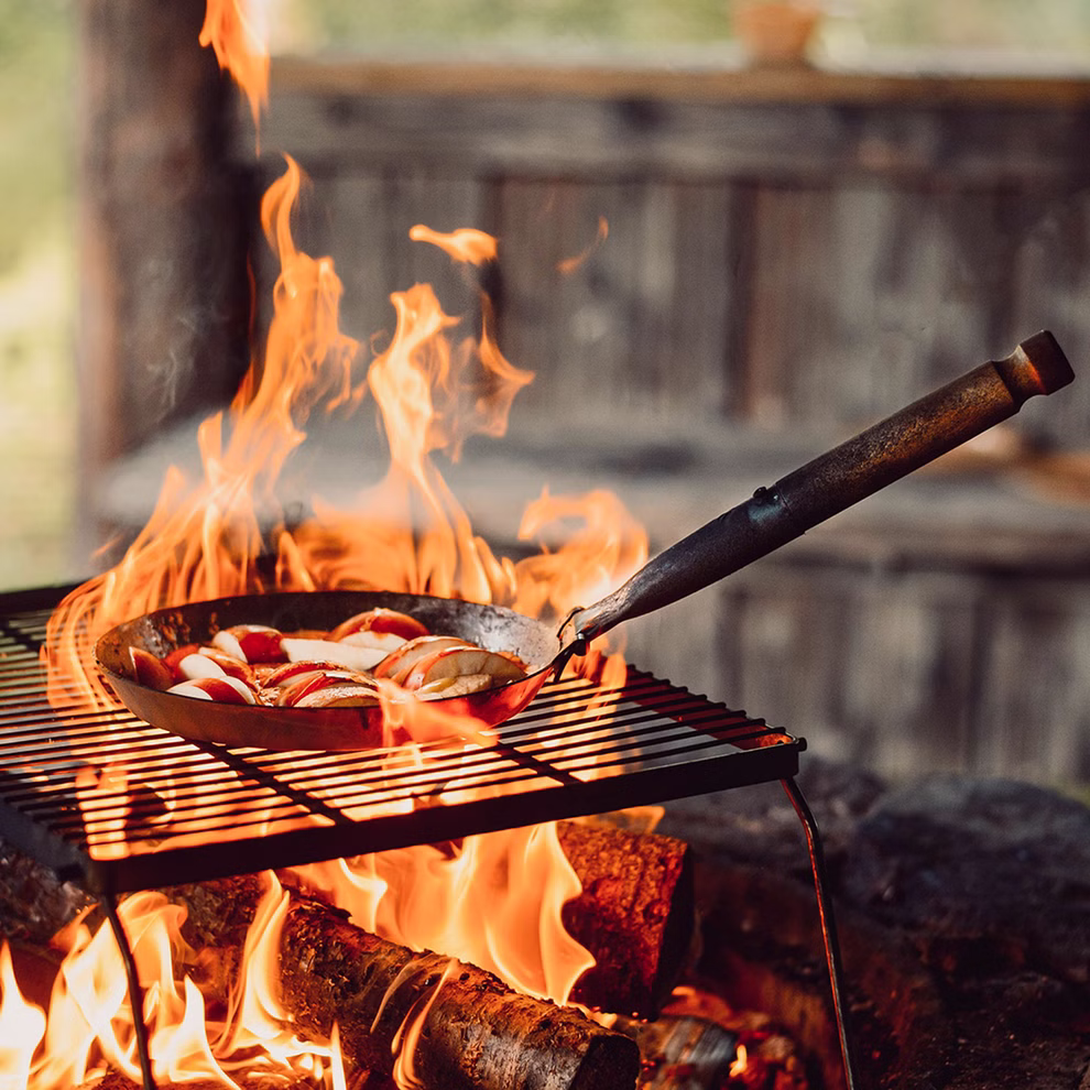 Stabilotherm Pfanne für das Lagerfeuer mit Klappgriff_31902 Die Stabilotherm Pfanne für das Lagerfeuer mit Klappgriff sitzt auf einem Grillrost über einem Holzfeuer gefüllt mit geschnittenem Gemüse. Ein Metallspatel ruht in der Pfanne die vor einem rustikalen Holzhintergrund steht.