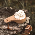 Stabilotherm Holzbecher 120 ml mit Elch Motiv _31910 Der Stabilotherm Holzbecher 120 ml mit Elch Motiv gefüllt mit Schlagsahne ruht auf einem Baumstumpf im Freien mit grünem Gras und Moos im Hintergrund.