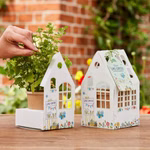 Die Stadtgärtner Gewächshäuschen Ellas Limo-Garten Anzucht-Set_32137 Eine Hand berührt eine getopfte Kräuterpflanze im Gewächshäuschen Ellas Limo-Garten Anzucht-Set von Die Stadtgärtner beide Sets in hausförmigen Halterungen mit bunten Motiven auf einem Holztisch im Freien.
