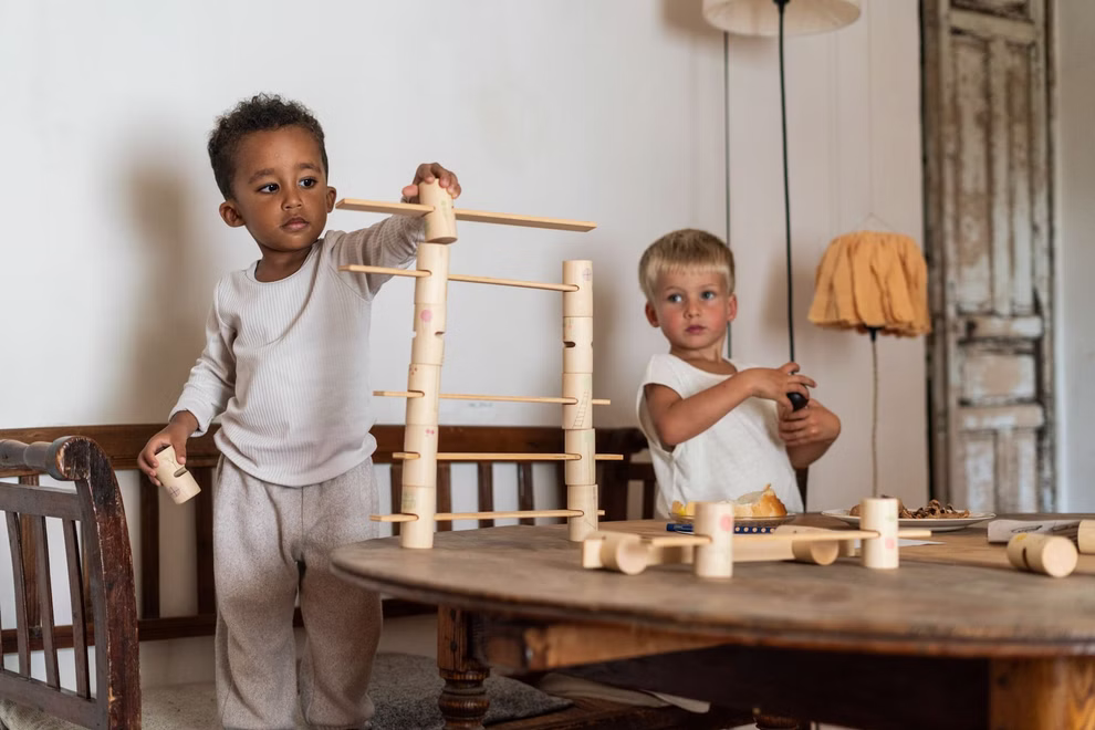 Grapat Grapat Woodland Konstruktionsspiel _32190 Zwei kleine Kinder spielen drinnen; eines baut mit dem Grapat Woodland Konstruktionsspiel von Grapat auf einer Bank während das andere in einem rustikalen gemütlichen Raum neben einem Tisch mit Spielzeug und Snacks steht.