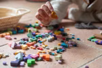 Grapat Grapat mis & match & bloom mit einem Bio-Baumwoll Beutel _32507 Eine Hand greift nach einer gelben Holzform aus dem Set mis & match & bloom von Grapat die zwischen anderen bunten Teilen auf einem gefliesten Boden verstreut liegt mit einer Aufbewahrungstasche aus Bio-Baumwolle in der Nähe.