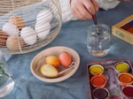 Eine Person taucht einen Pinsel in Wasser neben einer Palette mit Wasserfarben bemalten Grapat Sunrise Ostereiern (natur rot gelb) von Grapat und einem Korb mit unbemalten Eiern auf einem blauen Tischtuch.