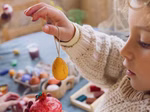 Ein kleines Kind in einem Strickpulli hält ein gelbes Grapat Sunrise Ostereier natur rot gelb von Grapat in die Höhe. Auf dem Tisch dahinter liegen weitere verzierte Eier und Bastelmaterial.