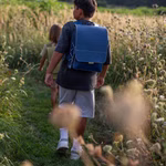 hukkepack Schulranzen Leder Blau Kompakt _32599 Zwei Kinder gehen im Freien auf einem überwucherten Weg umgeben von wilden Pflanzen und Sonnenlicht. Der Fokus liegt auf dem vorderen Kind das den hukkepack Schulranzen Leder Blau Kompakt Rucksack trägt.