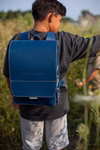 hukkepack Schulranzen Leder Blau Kompakt _32600 Ein Kind im Freien mit einem erhobenen Arm trägt den hukkepack Schulranzen Leder Blau Kompakt Rucksack mit sichtbaren Nähten und dem eingeprägten Markennamen. Im grasbewachsenen Hintergrund ist teilweise ein weiteres Kind zu sehen.