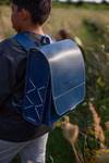 hukkepack Schulranzen Leder Blau Kompakt _32601 Ein Kind mit einem dunklen Hemd und einem großen blauen Hukkepack Schulranzen Leder Blau Kompakt steht auf einem grasbewachsenen Feldweg und schaut von der Kamera weg.