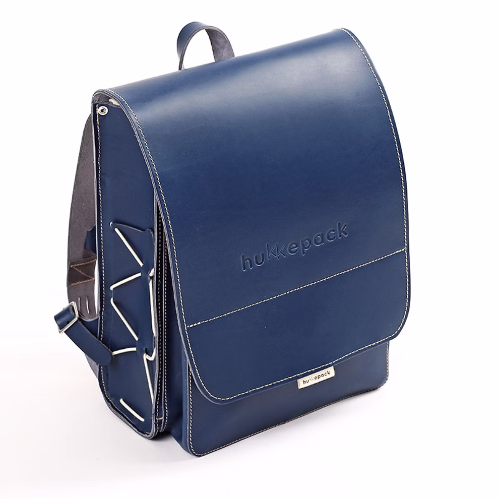 hukkepack Schulranzen Leder Blau Kompakt _32595 Der Schulranzen Leder Blau Kompakt von hukkepack ist ein blauer Lederrucksack mit weißen Nähten Tragegriff oben geprägtem Markenzeichen auf der Frontklappe und einem einzigartigen Zickzackmuster auf der linken Seite.