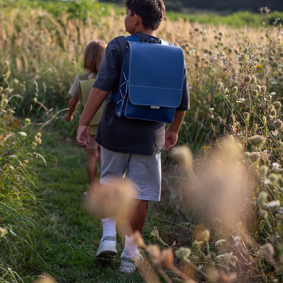 hukkepack Schulranzen Leder Blau Kompakt _32599 Zwei Kinder gehen im Freien auf einem überwucherten Weg umgeben von wilden Pflanzen und Sonnenlicht. Der Fokus liegt auf dem vorderen Kind das den hukkepack Schulranzen Leder Blau Kompakt Rucksack trägt.