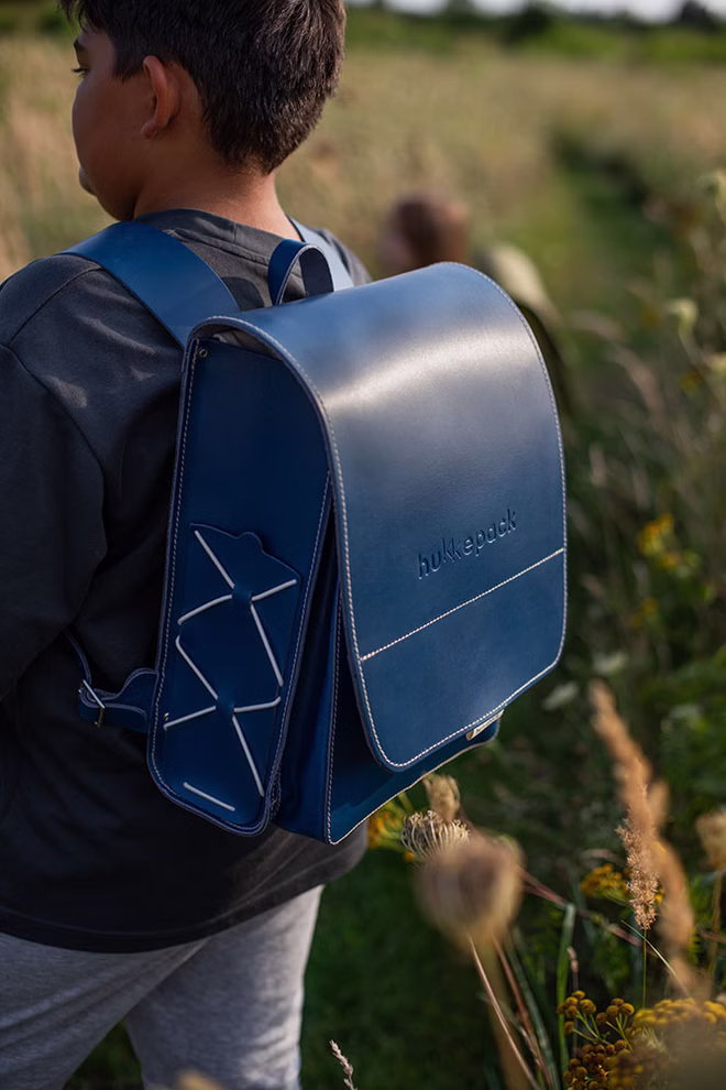 hukkepack Schulranzen Leder Blau Kompakt _32601 Ein Kind mit einem dunklen Hemd und einem großen blauen Hukkepack Schulranzen Leder Blau Kompakt steht auf einem grasbewachsenen Feldweg und schaut von der Kamera weg.