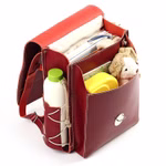 Der hukkepack Schulranzen Leder Rot Kompakt ist offen und bietet Platz für Bücher eine gelbe Lunchbox ein Stofftier eine Trinkflasche und eine Tasche mit Reißverschluss. Er ist mit weißen Nähten und einer Außenhalterung für Flaschen versehen.