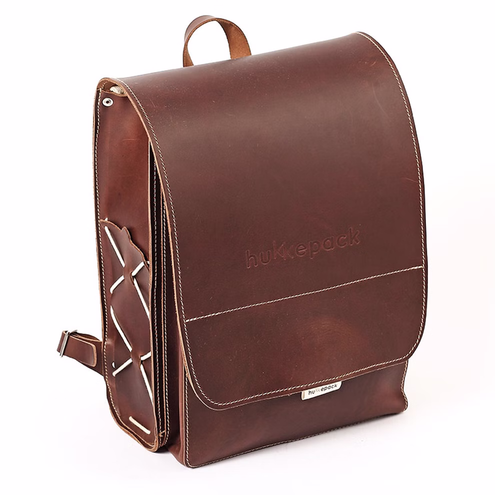 hukkepack Schulranzen Leder Braun Kompakt _32609 Der "Schulranzen Leder Braun Kompakt" von hukkepack ist ein brauner Lederrucksack mit weißen Nähten geprägtem Markennamen auf der Klappe und einer äußeren Seitentasche mit gekreuzten Riemen.