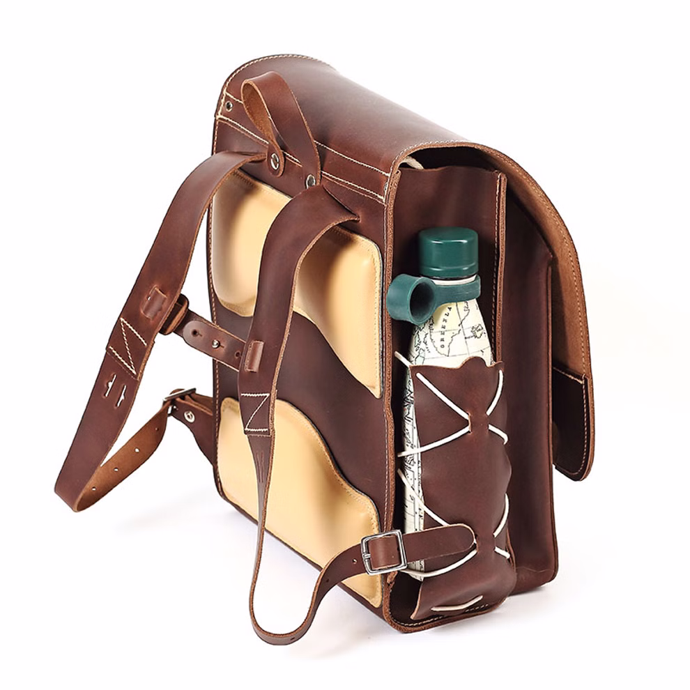 hukkepack Schulranzen Leder Braun Kompakt _32612 Der hukkepack Schulranzen Leder Braun Kompakt ist ein braun-hellbrauner Lederrucksack mit zwei Schulterriemen und einer externen Seitentasche in die eine mit einer Karte umwickelte und mit einer weißen Kordel gesicherte Wasserflasche mit grünem Deckel pass