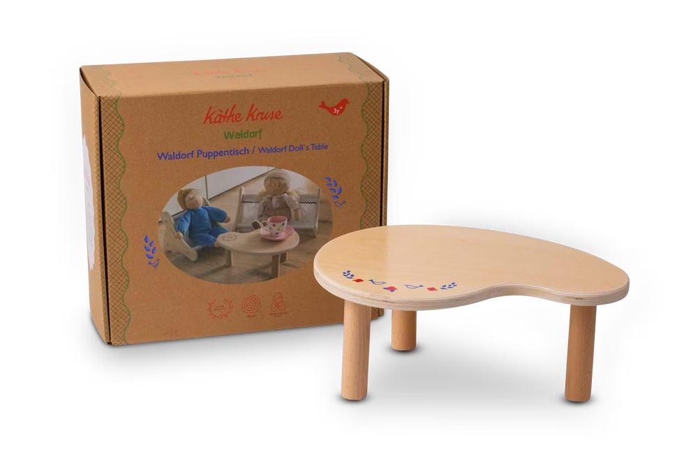 Käthe Kruse Waldorf Puppentisch Holz_33063 Ein kleiner leichter Waldorf Puppentisch Holz von Käthe Kruse mit drei Beinen wird neben seiner braunen Schachtel gezeigt auf der zwei Puppen am Tisch abgebildet und mit dem Marken- und Produktnamen beschriftet sind.
