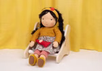 Eine handgefertigte Stoffpuppe mit lockigem schwarzem Haar sitzt auf einem Käthe Kruse Waldorf Puppenstuhl Holz trägt ein gelbes Stirnband einen senfgelben Pullover einen geblümten Rock und gestreifte Leggings vor einem gelben Vorhang.