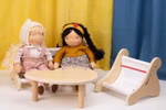 Käthe Kruse Waldorf Puppenbank Holz_33049 Zwei Käthe Kruse Waldorf Puppenbank Holz Stoffpuppen sitzen auf Holzstühlen an einem runden Tisch ein leerer Stuhl steht daneben im Hintergrund sind gelbe und blaue Vorhänge zu sehen.