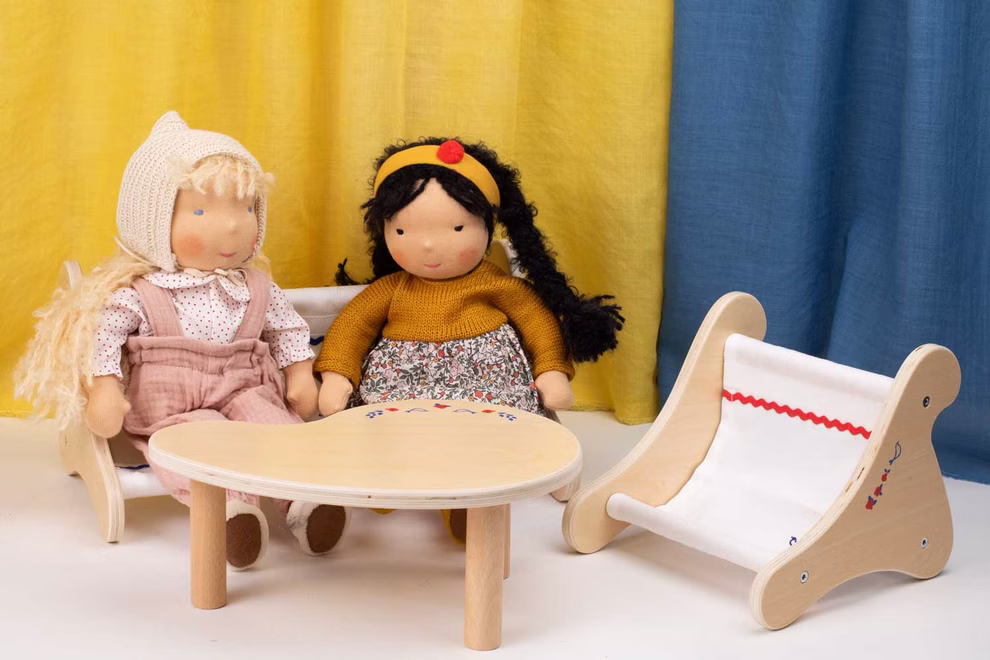 Käthe Kruse Waldorf Puppenbank Holz_33049 Zwei Käthe Kruse Waldorf Puppenbank Holz Stoffpuppen sitzen auf Holzstühlen an einem runden Tisch ein leerer Stuhl steht daneben im Hintergrund sind gelbe und blaue Vorhänge zu sehen.