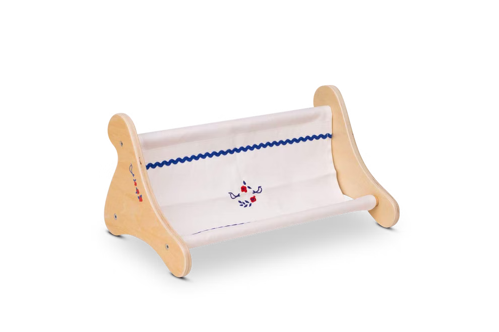 Käthe Kruse Waldorf Puppenbank Holz_33051 Die Käthe Kruse Waldorf Puppenbank Holz ist eine kleine hölzerne Puppenwiege mit geschwungenen Enden und Stoffseiten die mit blauen Verzierungen und rot-blau gestickten Blumenmustern versehen sind.