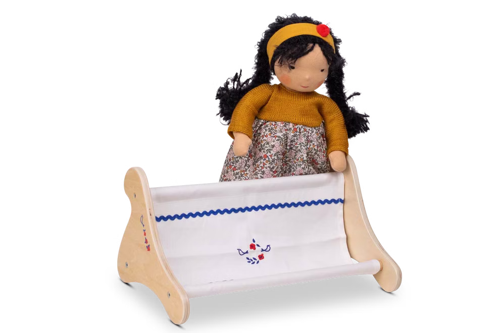 Käthe Kruse Waldorf Puppenbank Holz_33052 Vor einer Stoffpuppe mit langen dunklen Haaren gelbem Stirnband senfgelbem Pullover und geblümtem Rock sitzt ein Waldorf Puppenbank Holz Webstuhl von Käthe Kruse der teilweise weißen Stoff webt.