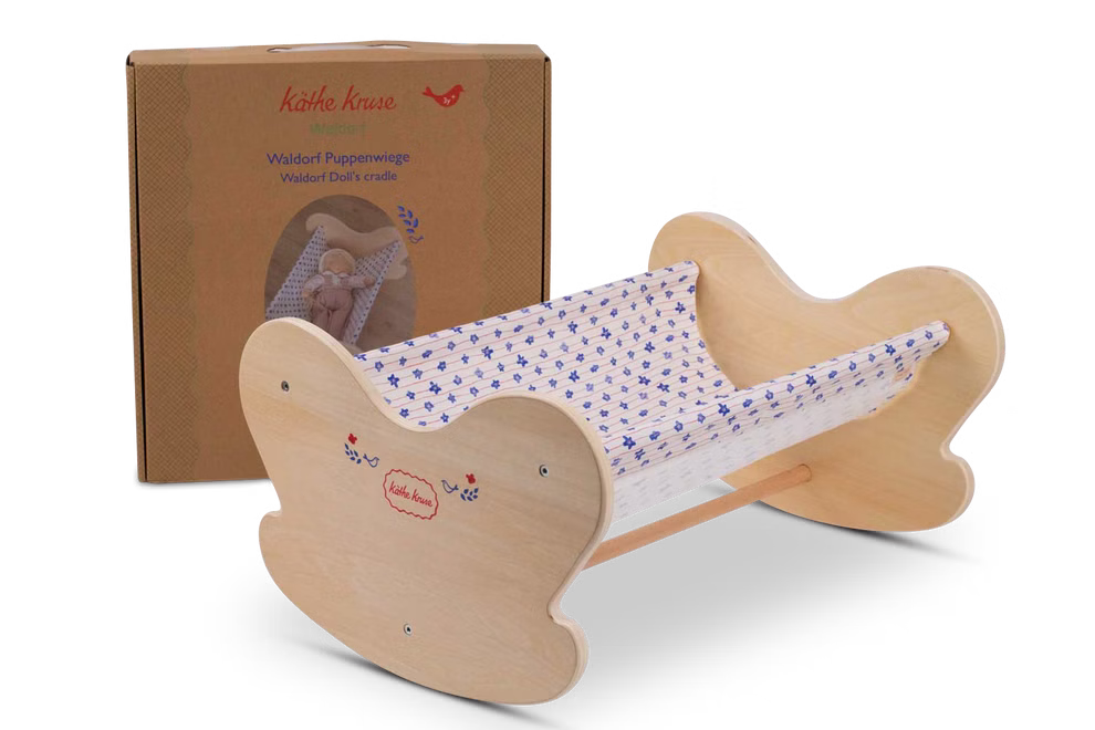 Käthe Kruse Waldorf Puppenwiege Holz_33046 Die Käthe Kruse Waldorf Puppenwiege Holz eine Holzpuppenwiege mit schmetterlingsförmigen Seiten und schlichtem Design ist mit weiß-blau gemustertem Bettzeug ausgestattet und wird vor dem braunen Käthe Kruse Karton präsentiert.