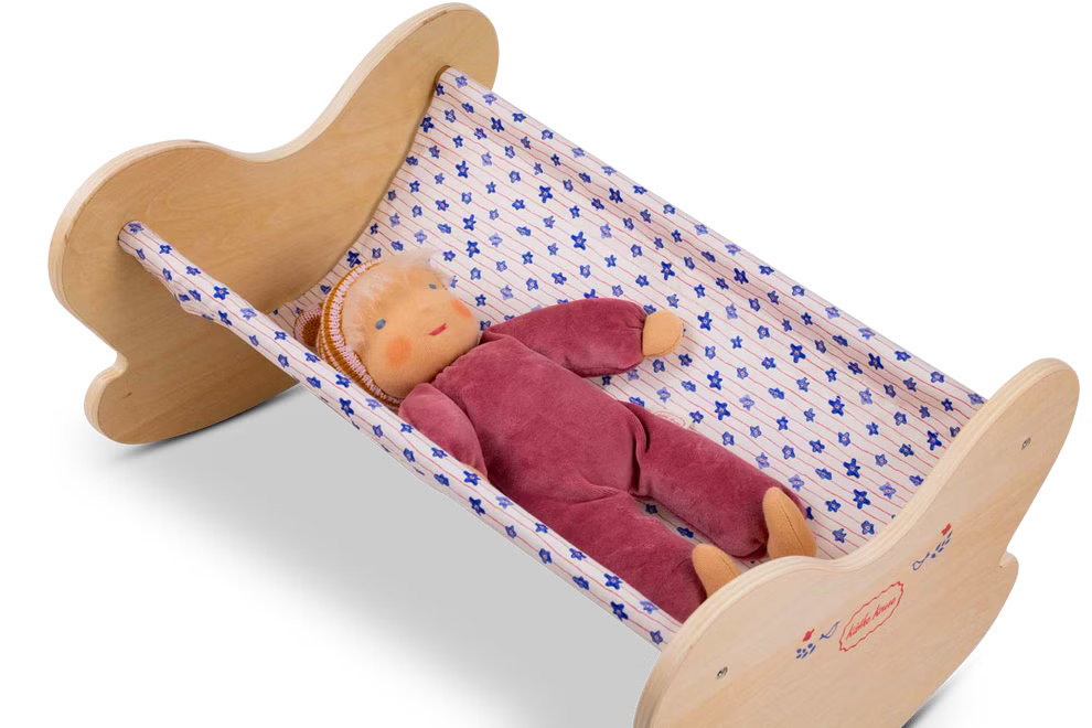 Käthe Kruse Waldorf Puppenwiege Holz_33047 Die Käthe Kruse Waldorf Puppenwiege Holz zeigt eine weiche Stoffpuppe in burgunderrotem Outfit und Strickmütze die in einer kleinen Holzwiege mit blau-weiß gemusterter Hängematte ruht.