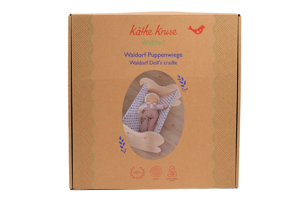 Käthe Kruse Waldorf Puppenwiege Holz_33048 Eine braune Pappschachtel mit blauen und grünen Akzenten zeigt das Set Käthe Kruse Waldorf Puppenwiege Holz mit einer kleinen Puppe in einer Holzwiege mit Kissen und Decke die durch ein Fenster auf der Verpackung sichtbar ist.