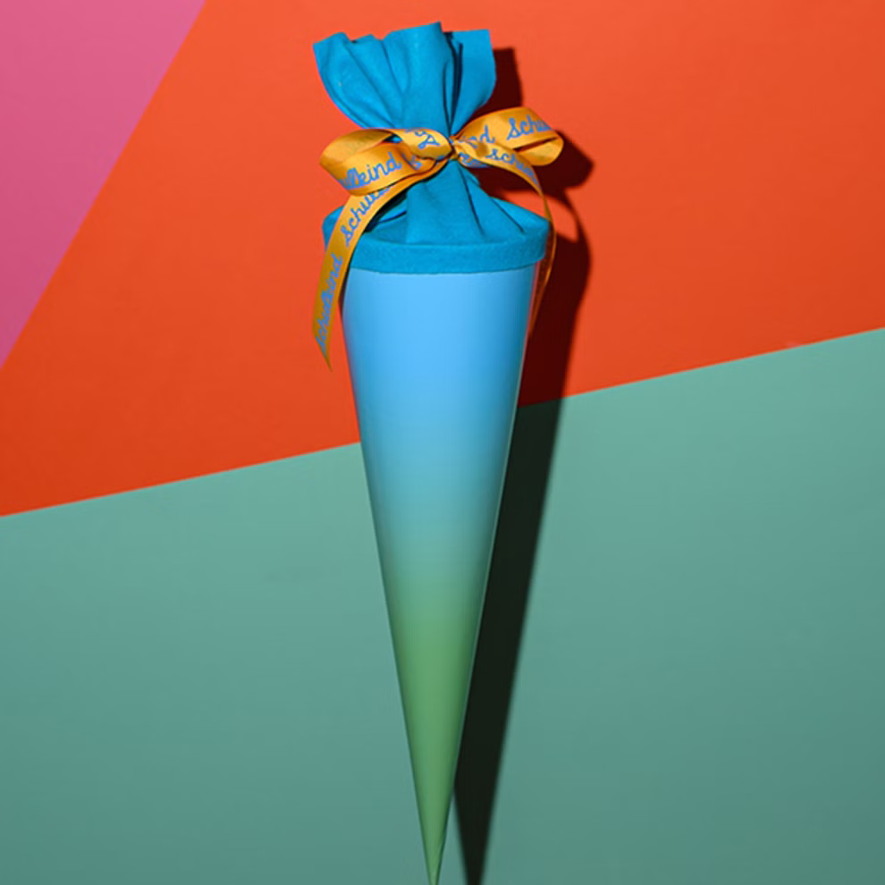 AVA & YVES Schultüte grün-türkis 35 cm_33319 Die Schultüte grün-türkis 35 cm von AVA & YVES ist eine kegelförmige Geschenktüte in Blau und Grün die mit einem gelben Band verschnürt ist und vor einem leuchtenden geometrischen Hintergrund in Orange Rosa und Grün steht.