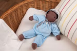 Waldorfkind Baby Puppe nach Waldorfart 46 cm_33708 Die Waldorfkind Baby Puppe nach Waldorfart 46 cm eine weiche braune Stoffpuppe in hellblauem Outfit und grau abgesetzter Haube liegt auf einem weißen Kissen in einem Weidenkorb neben einem gestreiften Kopfkissen.