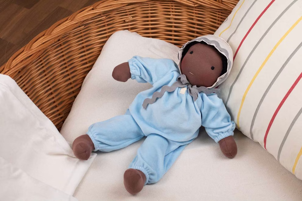 Waldorfkind Baby Puppe nach Waldorfart 46 cm_33708 Die Waldorfkind Baby Puppe nach Waldorfart 46 cm eine weiche braune Stoffpuppe in hellblauem Outfit und grau abgesetzter Haube liegt auf einem weißen Kissen in einem Weidenkorb neben einem gestreiften Kopfkissen.