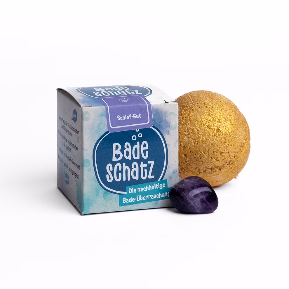 BadeSchatz Schlaf Gut Badebombe mit Goldstaub und Edelstein Amethyst neben der Papierverpackung.