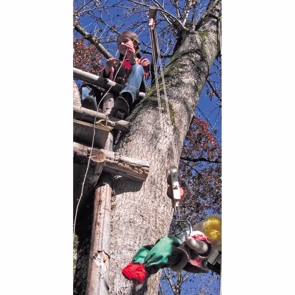 Kraul Bausatz Flaschenzüge _1507 Ein Kind sitzt auf einer hölzernen Baumhausplattform und benutzt das Flaschenzugsystem von Kraul Bausatz um ein bunt gekleidetes Spielzeug vor einem strahlend blauen Himmel auf den Baum zu heben.