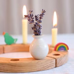 Die mit Lavendelzweigen gefüllte Grimms Stecker Vase ruht auf einem Holzständer und ist von zwei brennenden Kerzen und lebhaften Holzdekorationen wie einem Regenbogen und einem Vogel umgeben. Der sanft verschwommene Hintergrund verleiht ihr eine zarte Note