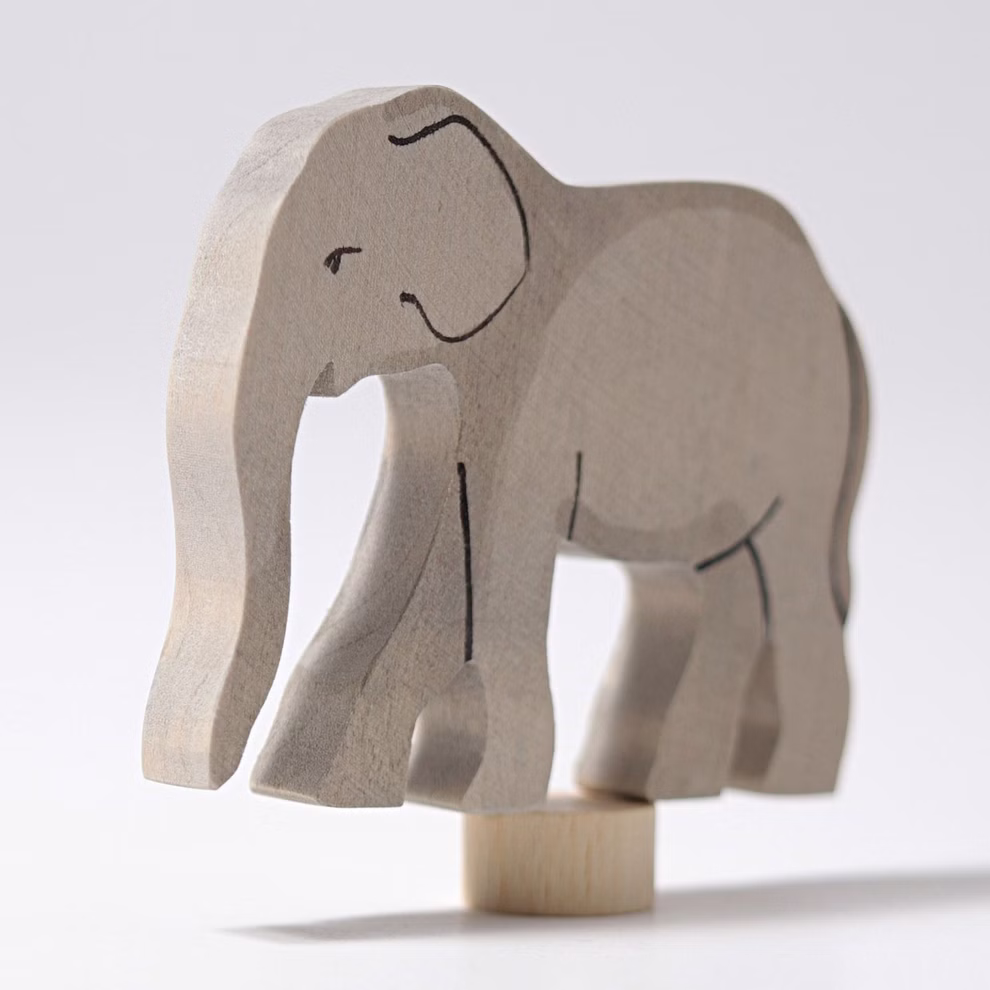 Der Grimms Geburtstagsstecker Elefant von Grimm's ist ein einfach geschnitzter hölzerner Spielzeugelefant der mit minimalen schwarzen Linien für Details bemalt ist und auf einem kleinen Holzzylinder vor einem schlichten weißen Hintergrund balanciert.