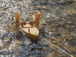 Kraul Forelle - Schiffsbausatz Bausatz_14326 Der Kraul Forelle - Schiffsbausatz Bausatz ist ein kleiner handgefertigter Holzboot-Bausatz mit Spule und Paddeln der an einer Schnur befestigt auf dem sonnenbeschienenen flachen Wasser schwimmt.
