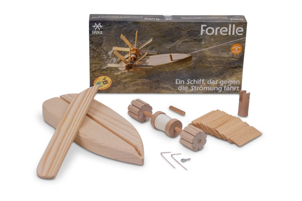 Kraul Forelle - Schiffsbausatz Bausatz_31639 Der Kraul Forelle - Schiffsbausatz Bausatz mit montierten Holzteilen Werkzeug und deutscher Anleitung ist vor seiner Schachtel mit der Aufschrift "Forelle" abgebildet; im Hintergrund schwimmt das Boot auf dem Wasser.