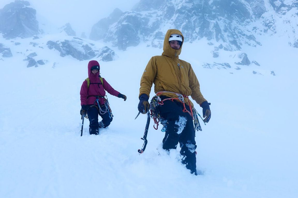 Zwei Winterkletterer in Isolationsjacken mit Kapuze stapfen mit Klettermaterial und Eisger&auml;ten durch tiefen Schnee