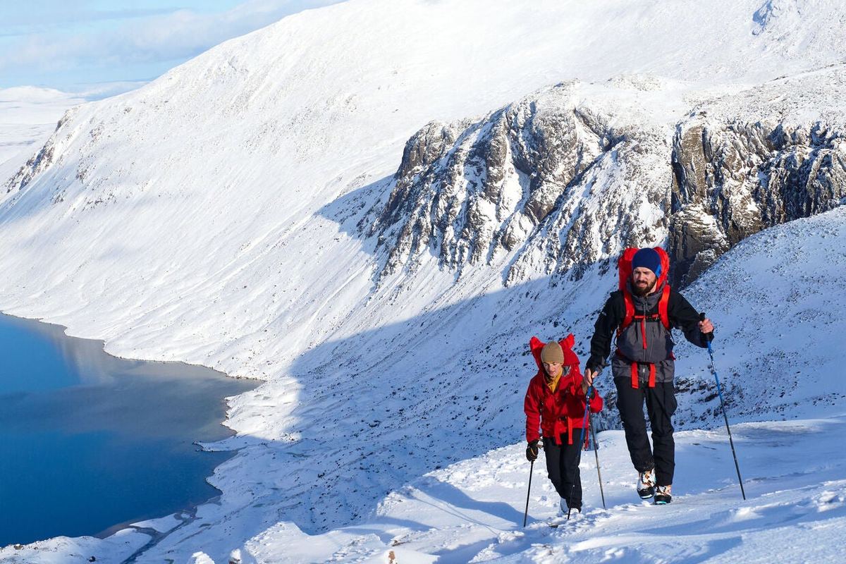 Zwei Wanderer mit St&ouml;cken und Rucks&auml;cken steigen an einem verschneiten Hang &uuml;ber einem See auf