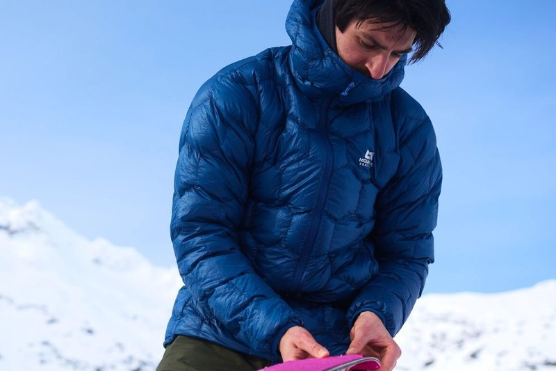 Skitourengeher in einer blauen Daunenjacke mit sechseckigen Steppkammern hantiert vor verschneiten Bergen mit seinem Steigfell