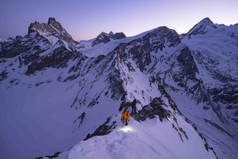 Bergsteiger mit Stirnlampe und oranger Daunenjacke auf einem verschneiten Grat in der D&auml;mmerung