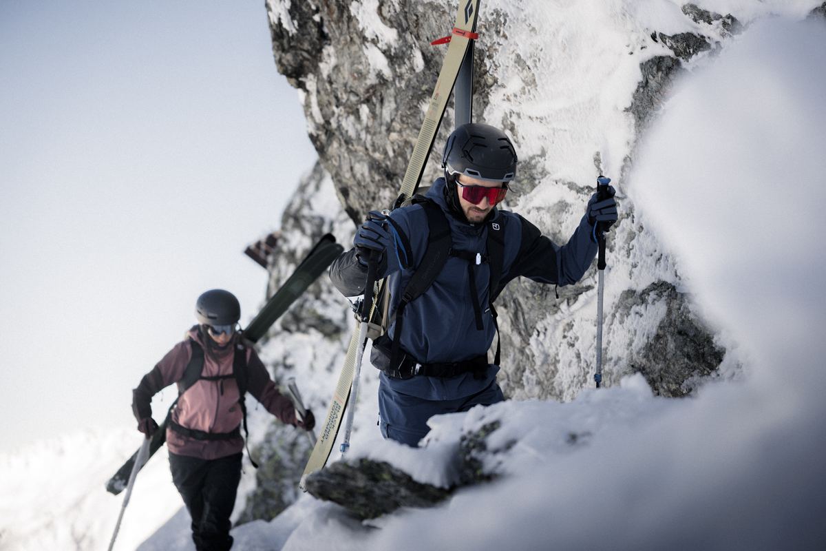 Zwei Skitourengeher mit Ski am Rucksack steigen durch eine verschneite Felsrinne auf