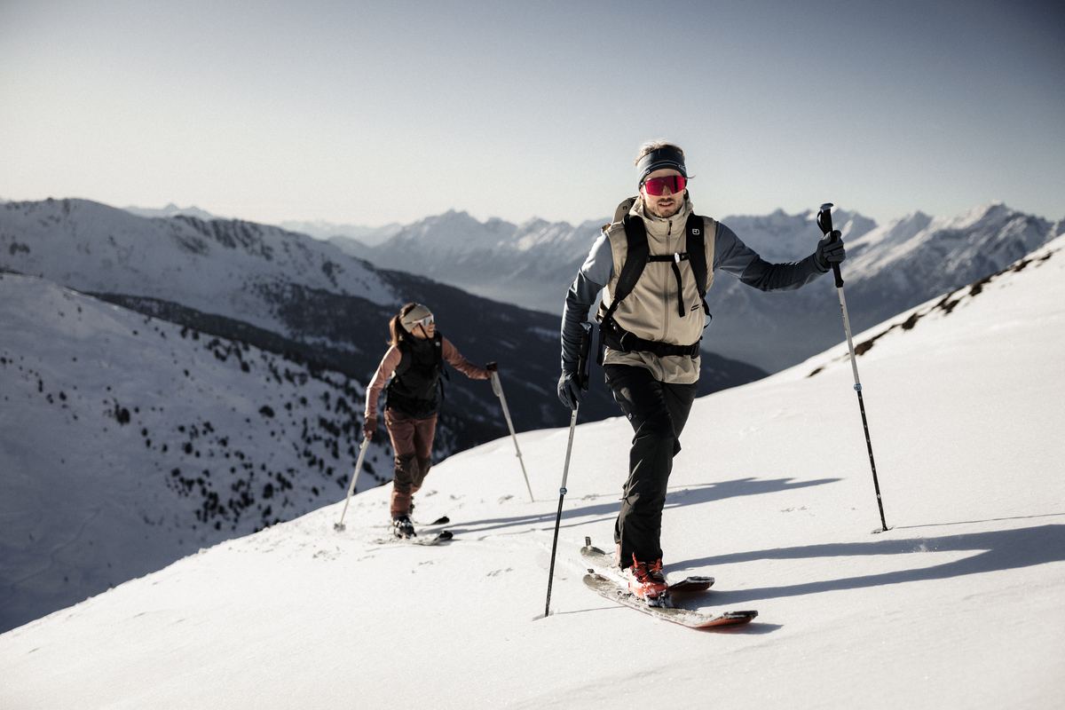 Skitourengeherin und Skitourengeher im Aufstieg mit Fellen vor einem Bergpanorama