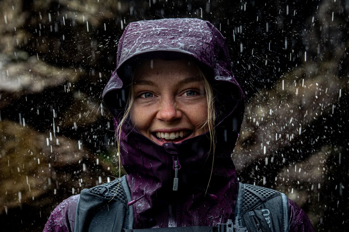 Lachende Wanderin mit Rucksack und aufgesetzter Kapuze einer violetten Hardshelljacke im Regen