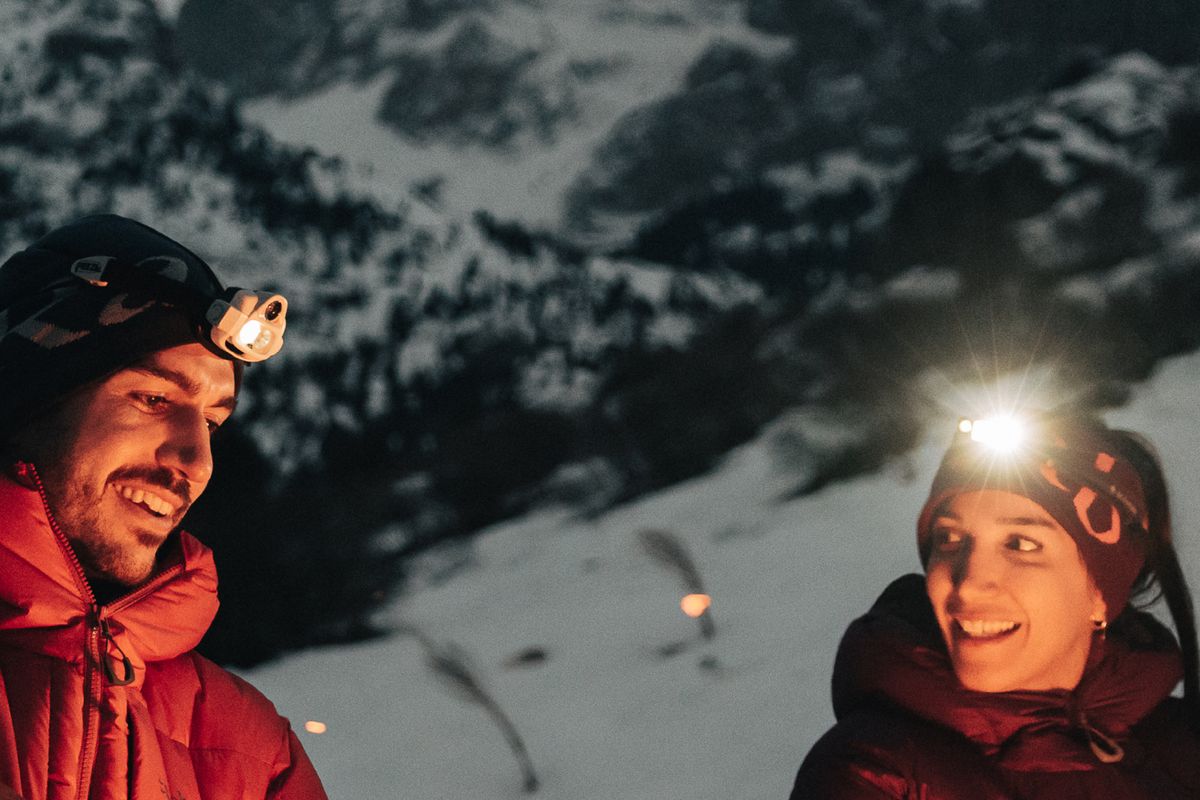 Zwei Personen mit Stirnlampen und Daunenjacken lachen abends vor verschneiten Bergen