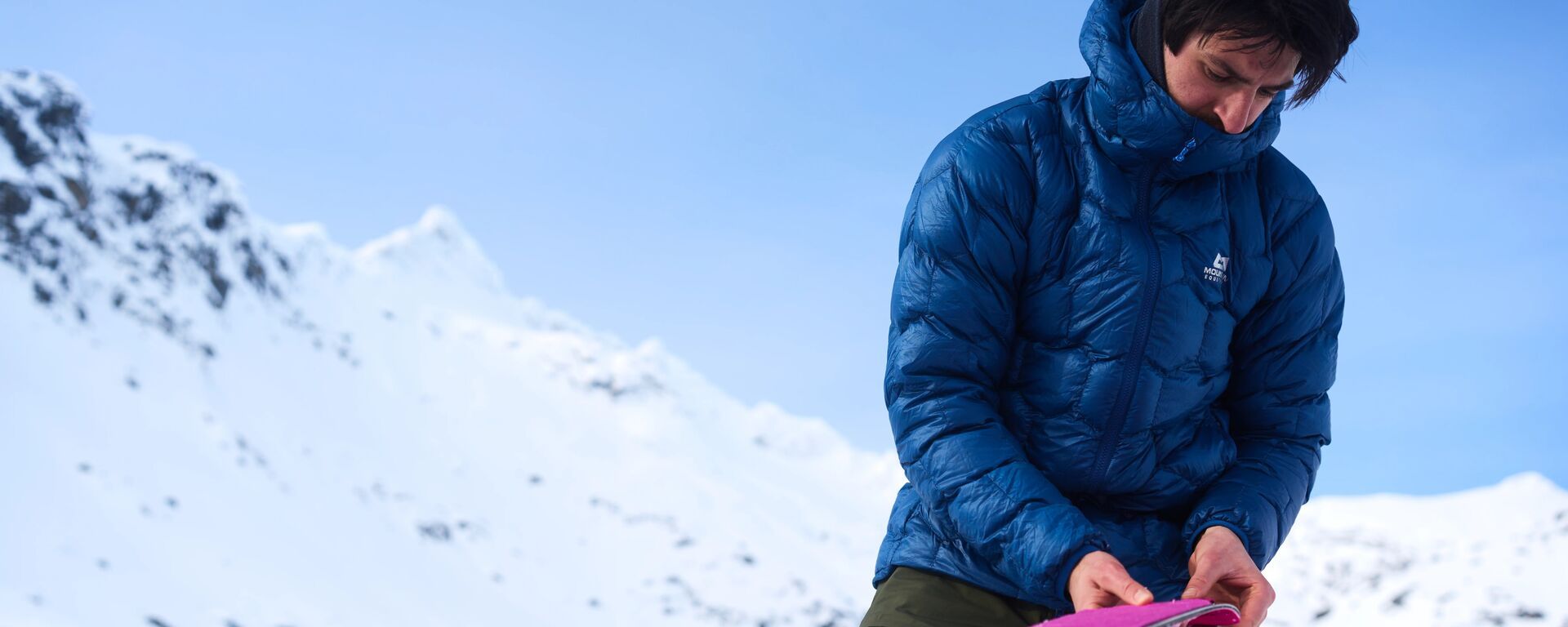 Skitourengeher in einer blauen Daunenjacke mit sechseckigen Steppkammern hantiert vor verschneiten Bergen mit seinem Steigfell