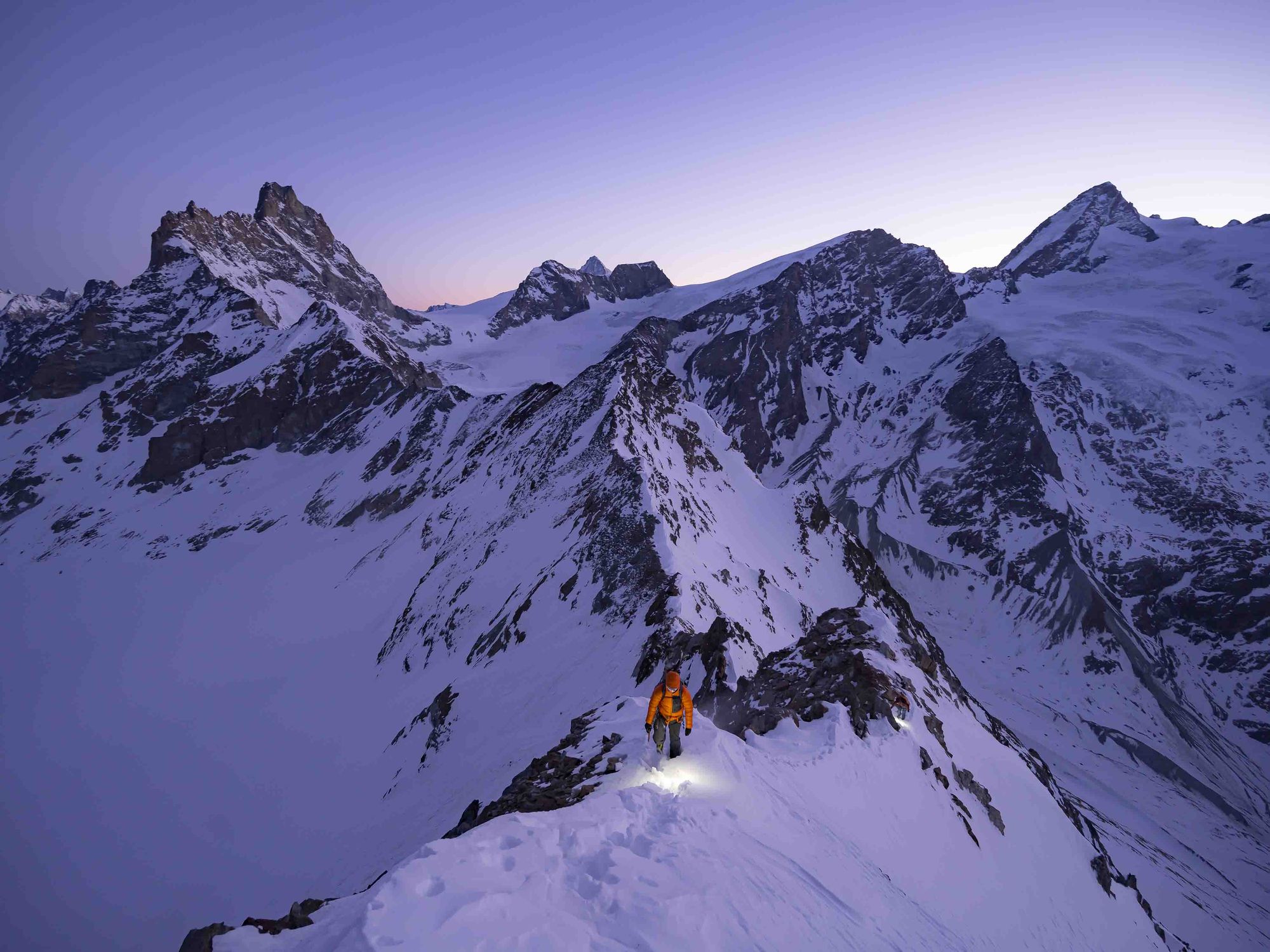 Bergsteiger mit Stirnlampe und oranger Daunenjacke auf einem verschneiten Grat in der D&auml;mmerung