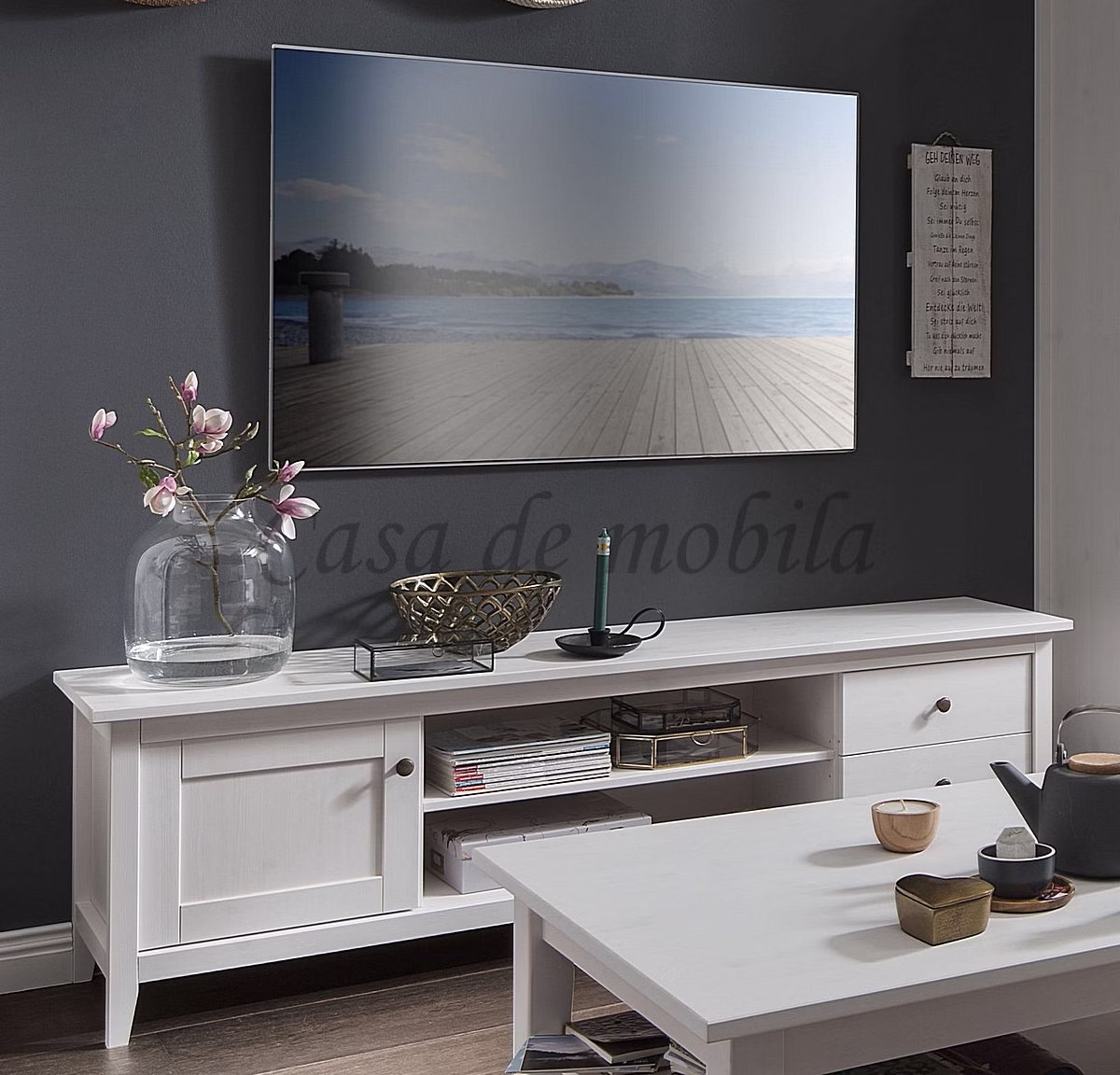 SOLVITA – TV-Lowboard im Wohnzimmer Weißes TV-Lowboard mit Fernseher vor dunkler Wand und Dekoration auf der Deckplatte.